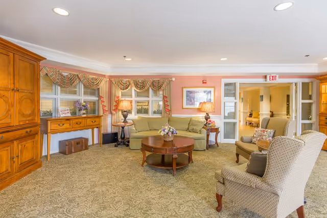 Communal sitting area with sofas, armchairs, a round coffee table, wooden cabinets, and lamps in a senior living facility.