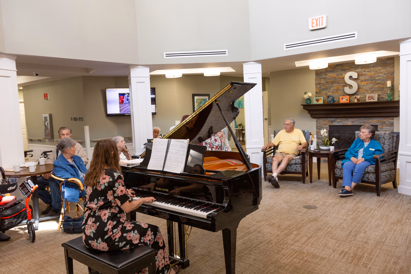 A woman in a floral dress plays a grand piano in a senior living facility common area. Several elderly residents sit around the room, listening and watching. The room has beige carpet, a stone fireplace with a large letter 'S' above it, and comfortable chairs arranged in a semi-circle. There is an exit sign above a doorway in the background.