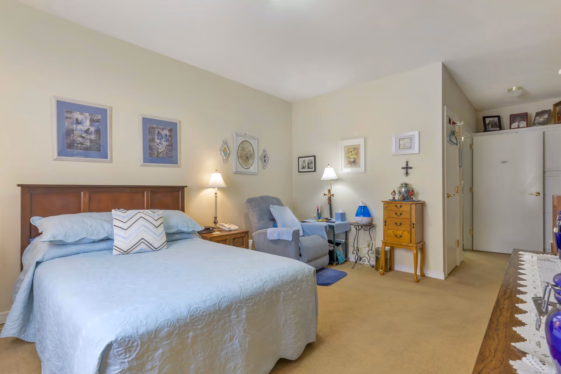A cozy bedroom in an assisted living facility featuring a bed with light blue bedding and a decorative pillow, a wooden headboard, two bedside tables with lamps, a comfortable recliner chair with a side table, framed artwork on the walls, and a small wooden dresser. The room has beige carpet and light-colored walls.