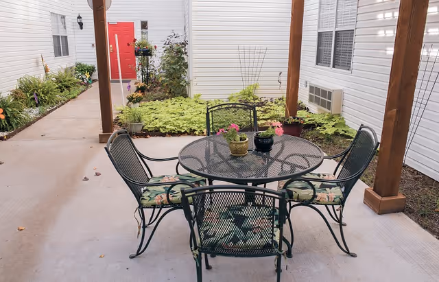 Outdoor patio area with a round metal table and four metal chairs with floral cushions. The patio is covered by a wooden pergola and surrounded by garden beds with various plants and flowers. A red door is visible in the background on a white building.