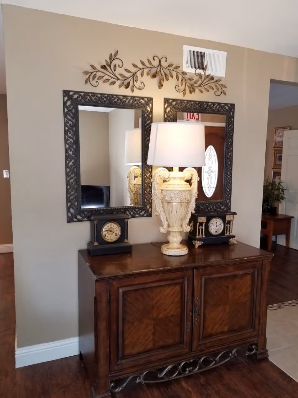 Wooden console table topped with an ornate urn-style lamp, two clocks, and reflected mirrors beneath decorative wall art in an interior entryway.