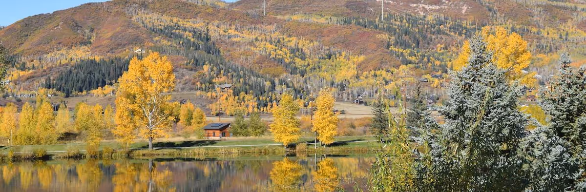 Scenic lakeside view showing autumn-colored trees and a small cabin reflected in calm water with forested hills in the background.