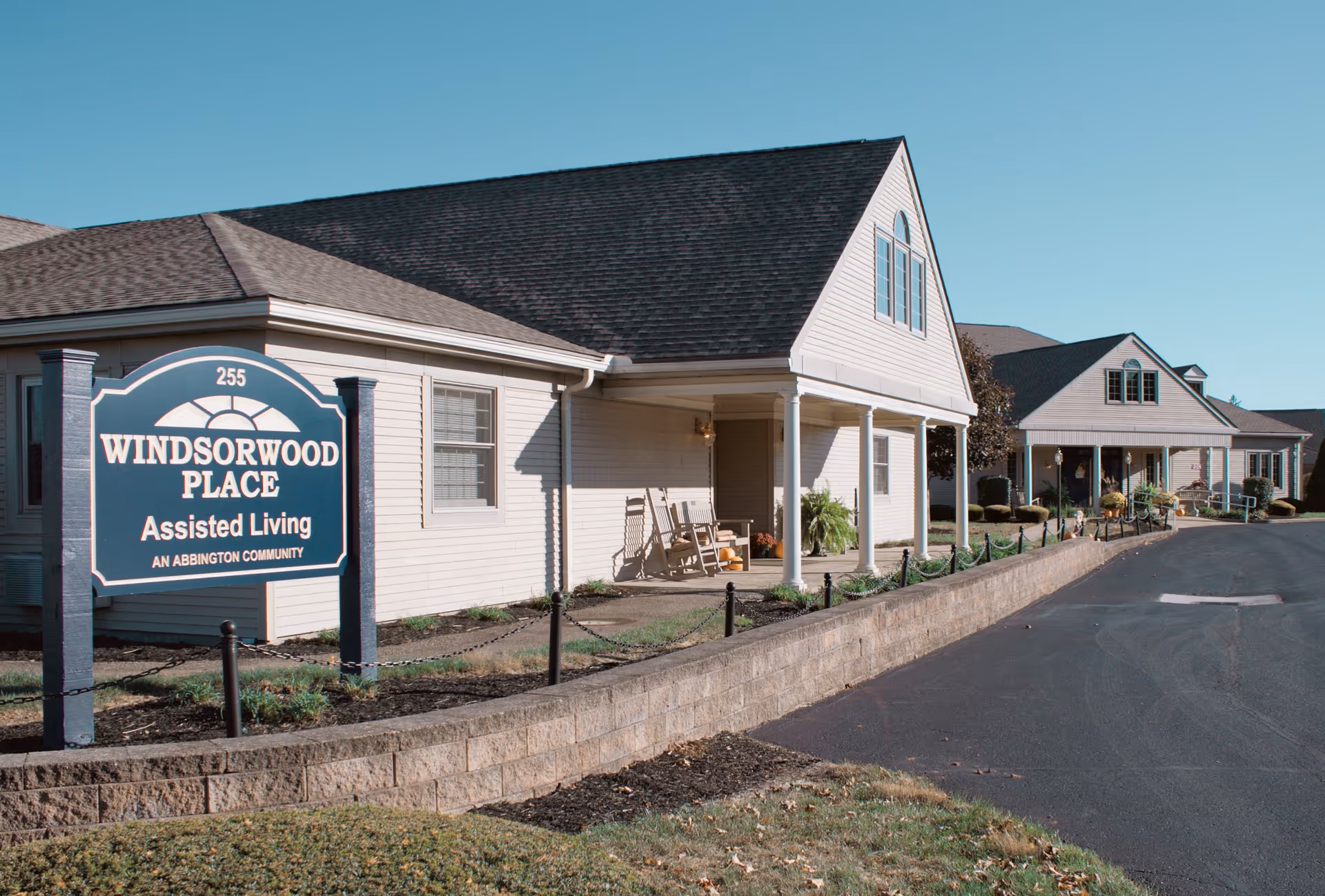 Exterior view of Windsorwood Place Assisted Living community buildings with a clear blue sky. The buildings have light-colored siding, dark roofs, and a covered porch with rocking chairs. A sign in front reads 'Windsorwood Place Assisted Living An Abbington Community'.