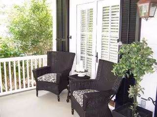 A porch area with two dark wicker chairs with patterned cushions, a small round table between them holding a decorative item, white railing, a potted plant, and white French doors with shutters leading inside.