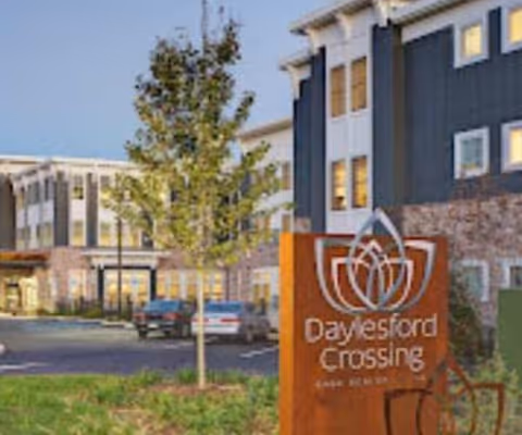 Exterior view of the Daylesford Crossing senior living building with its wooden sign in the foreground and parking/entrance behind.