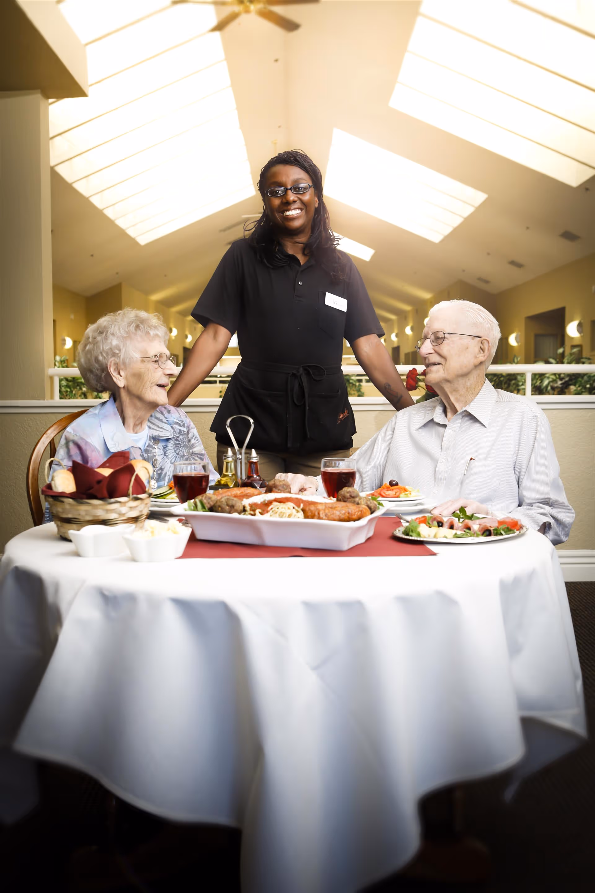 A smiling waitress stands behind an elderly man and woman seated at a round dining table with a white tablecloth. The table is set with plates of food, glasses of red beverage, a basket of bread, and condiments. The setting appears to be a well-lit dining area with a high ceiling and skylights.