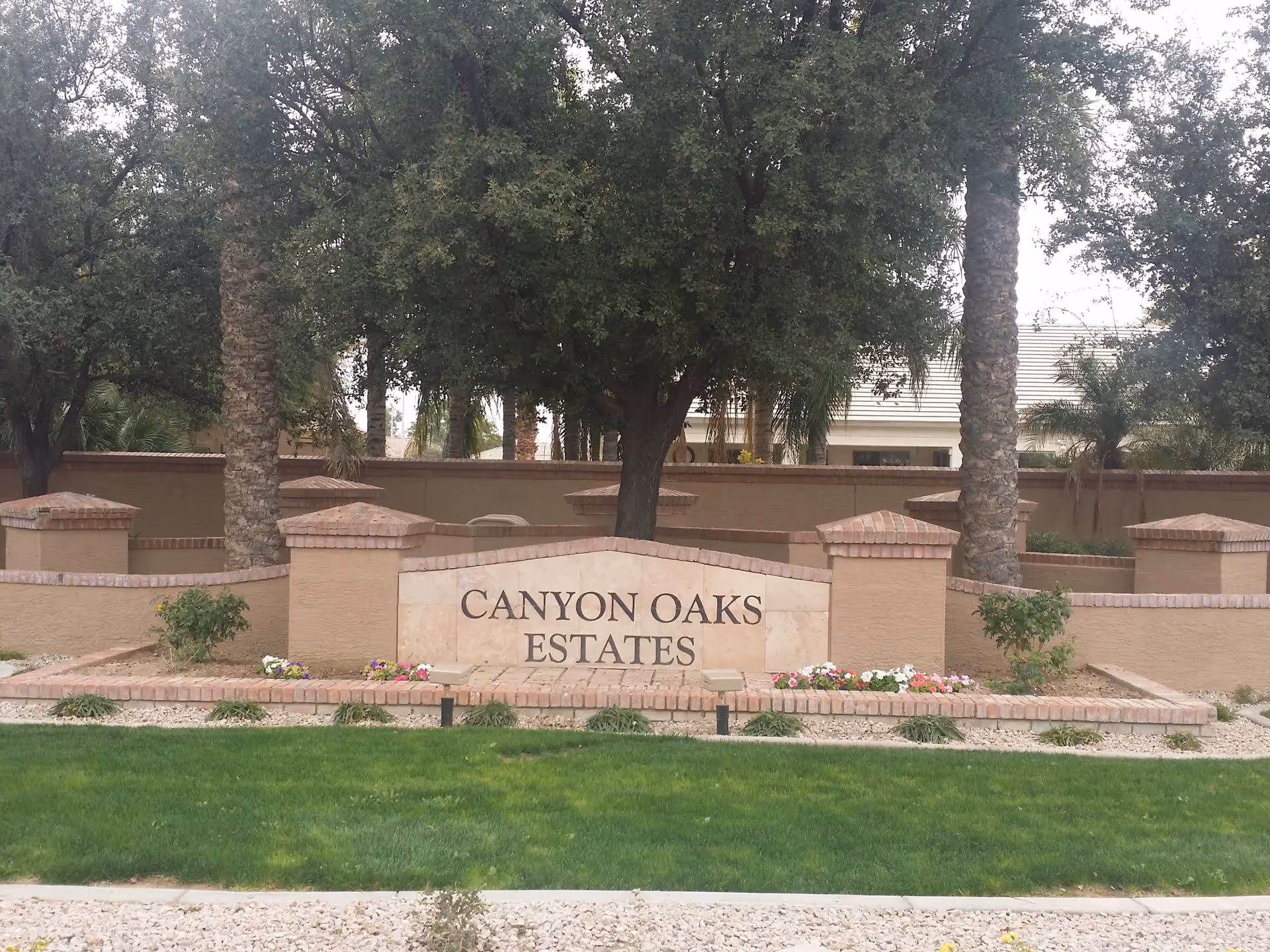 Stone entrance sign reading 'Canyon Oaks Estates' in front of trees, palms, and a low stucco wall.