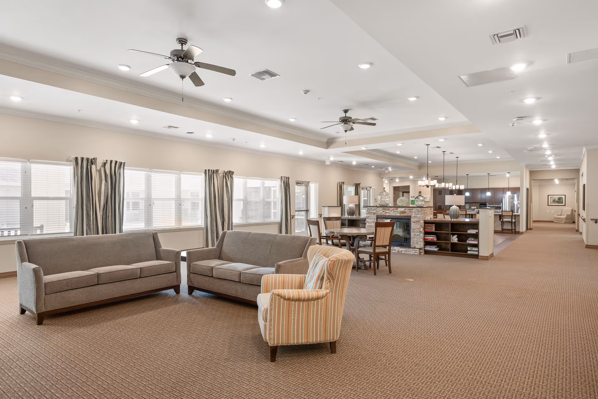 Spacious senior living common area with sofas and an armchair facing a fireplace, dining table and an open kitchen in the background.