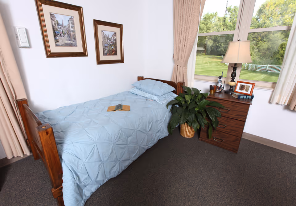 Small single bedroom with a wooden bed dressed in a light blue comforter, a nightstand with a lamp and plant, and a large window overlooking a green lawn.