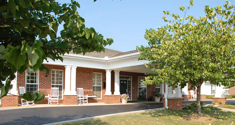 Exterior view of a single-story brick building with white columns and several windows. There are white rocking chairs on the porch and a tree with green leaves in the foreground under a clear blue sky.