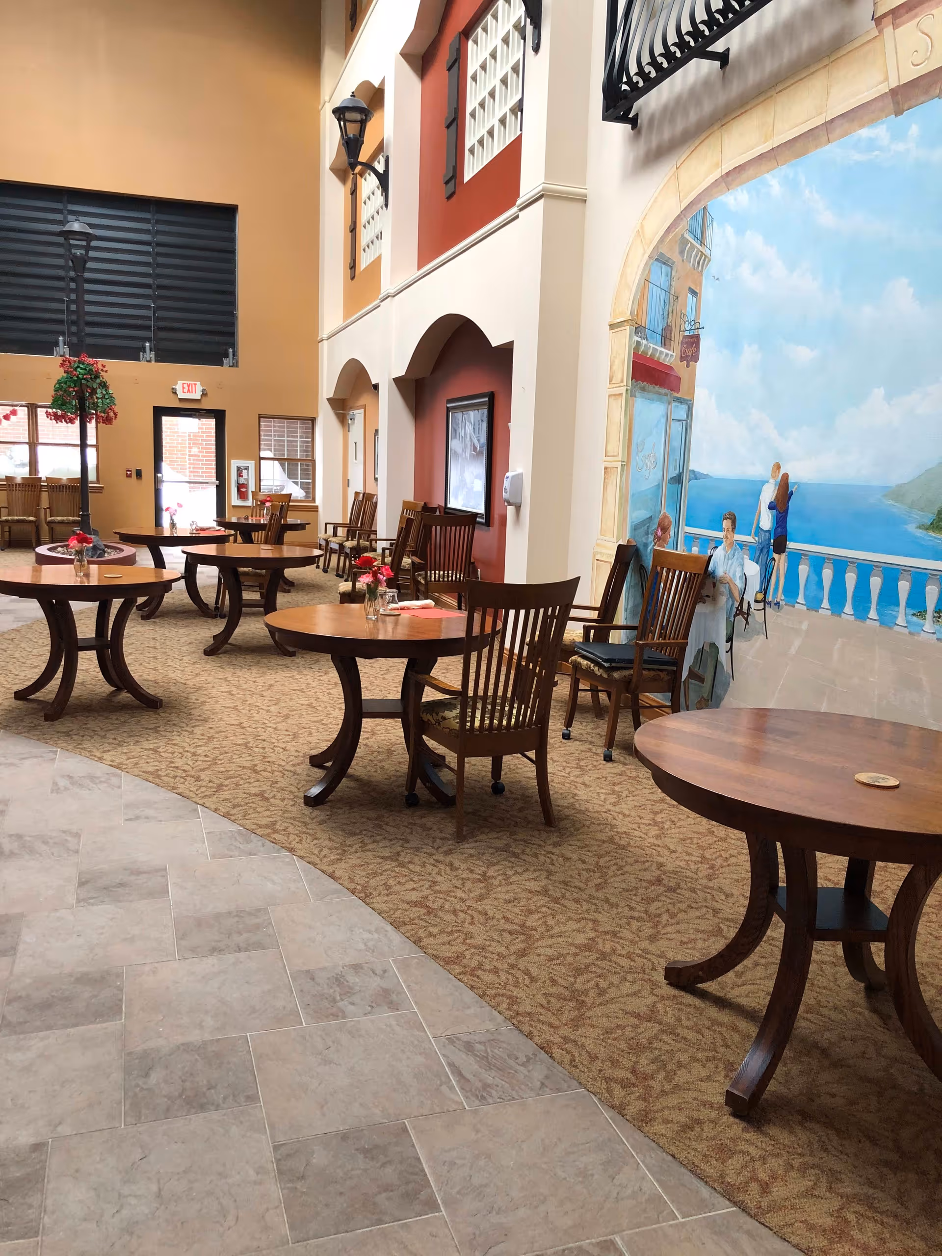 Interior view of a common area in Sherwood Lodge Assisted Living featuring round wooden tables with chairs on a carpeted floor. The walls have a warm color palette with a large mural depicting a seaside scene with people sitting and standing on a balcony overlooking the ocean. There are decorative streetlamp-style lights and an exit door in the background.