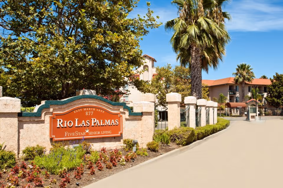 Entrance sign for Rio Las Palmas Five Star Senior Living with a stone wall and landscaped flowers in front, palm trees and other greenery, and a driveway leading to the senior living facility buildings under a clear blue sky.