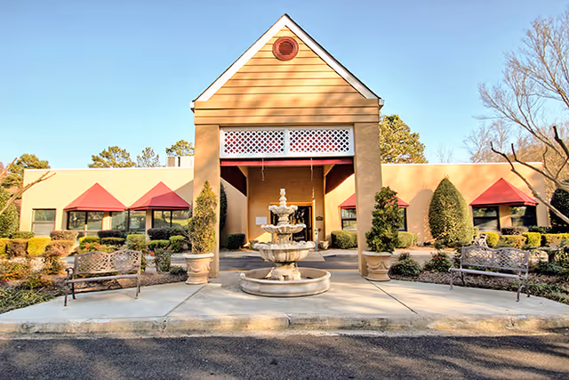 Front exterior view of Brookdale Charlotte East facility featuring a beige building with a peaked roof entrance, red awnings over windows, a central three-tiered water fountain, benches on either side, and landscaped bushes and trees.