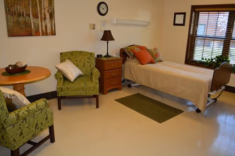 A senior living facility room with a single bed covered in beige bedding, two green upholstered armchairs with patterned fabric, a small wooden round table with a decorative bowl, a wooden nightstand with a lamp, a wall clock, a framed picture, and a window with wooden blinds.