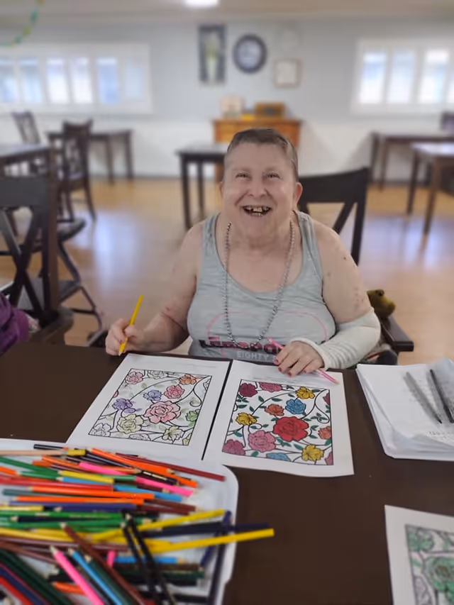 An elderly woman sitting at a table in a communal room, smiling while coloring floral designs on paper with colored pencils. The room has wooden floors, several tables and chairs, and windows letting in natural light.