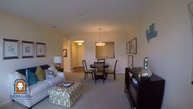 Interior view of a senior living facility's common area featuring a beige sofa with colorful cushions, a patterned ottoman, a dining table with four chairs, framed artwork on the walls, and a dark wooden cabinet with decorative items. The space is well-lit with ceiling lights and has a neutral color scheme.