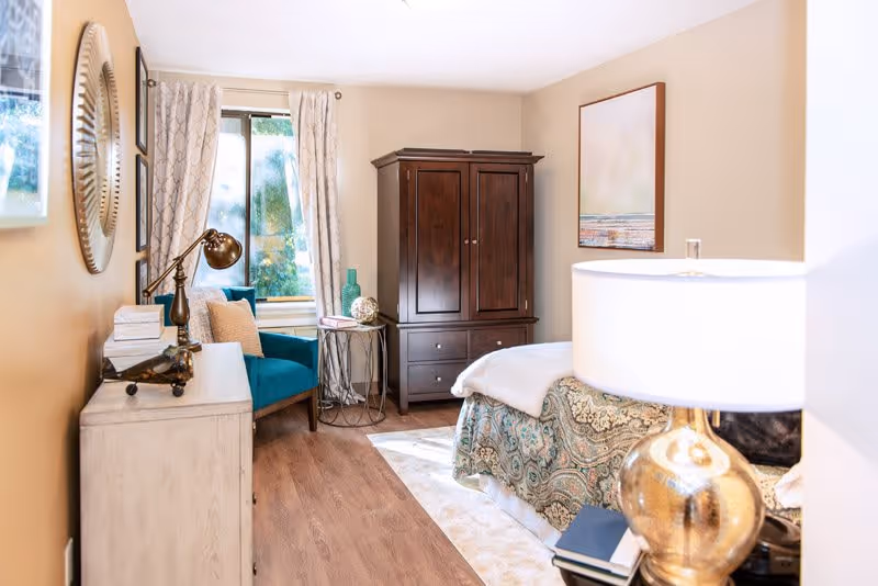 A cozy bedroom with a bed covered in a patterned bedspread, a wooden armoire, a blue armchair next to a window with patterned curtains, a small round side table with decorative items, a dresser with a lamp and decor, and a framed artwork on the wall.