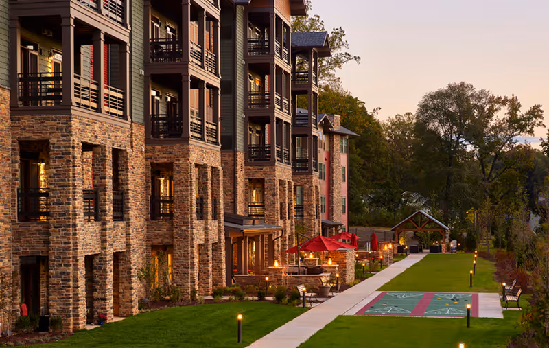 Exterior view of a multi-story senior living facility with stone and wood siding, balconies, a well-maintained lawn, a paved walkway, outdoor seating areas with red umbrellas, and shuffleboard courts, surrounded by trees at sunset.