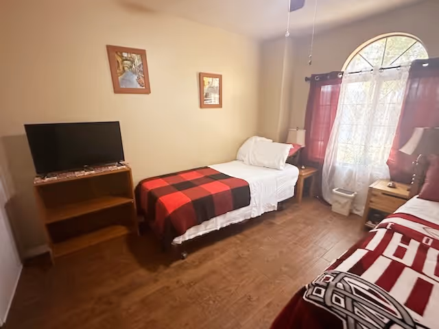 A small bedroom with two single beds, one with a red and black checkered blanket and the other with a red and white patterned blanket. There is a wooden TV stand with a flat-screen TV on the left side of the room. The room has wooden flooring, beige walls, two framed pictures on the wall, and a window with red curtains and white sheer curtains letting in natural light. There are two bedside tables with lamps next to each bed.