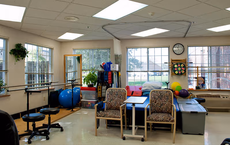 A well-lit therapy or exercise room with large windows letting in natural light. The room contains two patterned armchairs with a small table between them, exercise balls, stools on wheels, resistance bands, and various therapy equipment. There are ballet barres along the walls and a clock above a dartboard on the wall. The room has a clean and organized appearance.