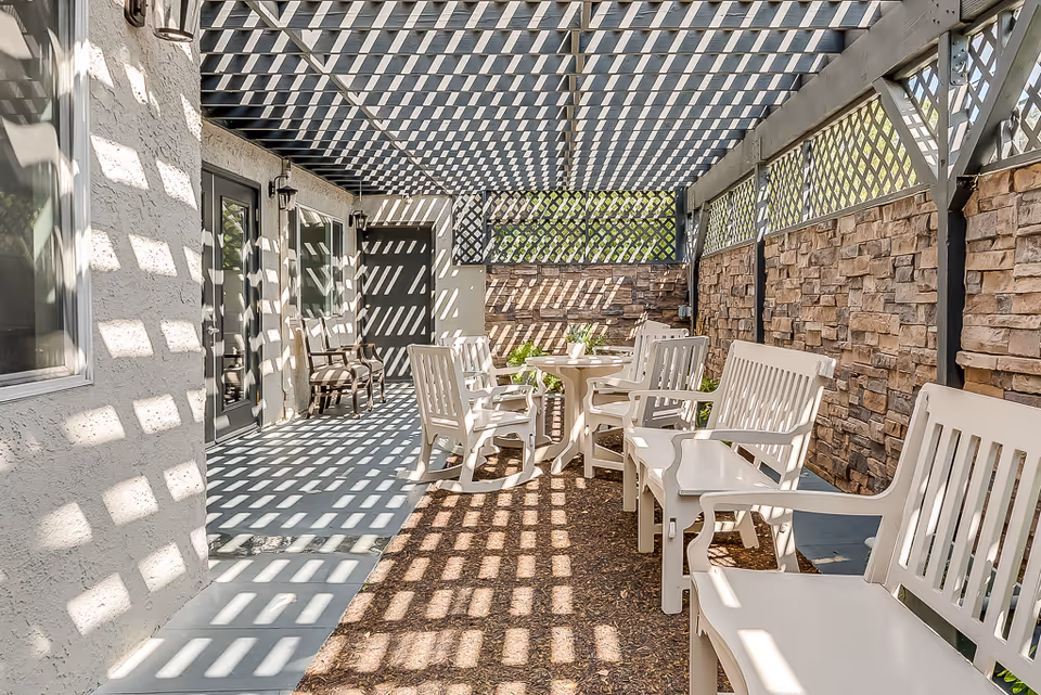 Outdoor patio area with white chairs and tables under a pergola casting a checkered shadow pattern. The space is enclosed by a stone wall and lattice panels, with doors and windows on one side.
