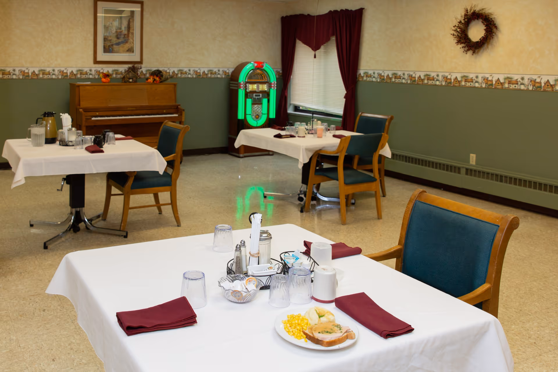 Dining room with white-tablecloth tables set for a meal, wooden chairs, a piano and a glowing jukebox against the wall.