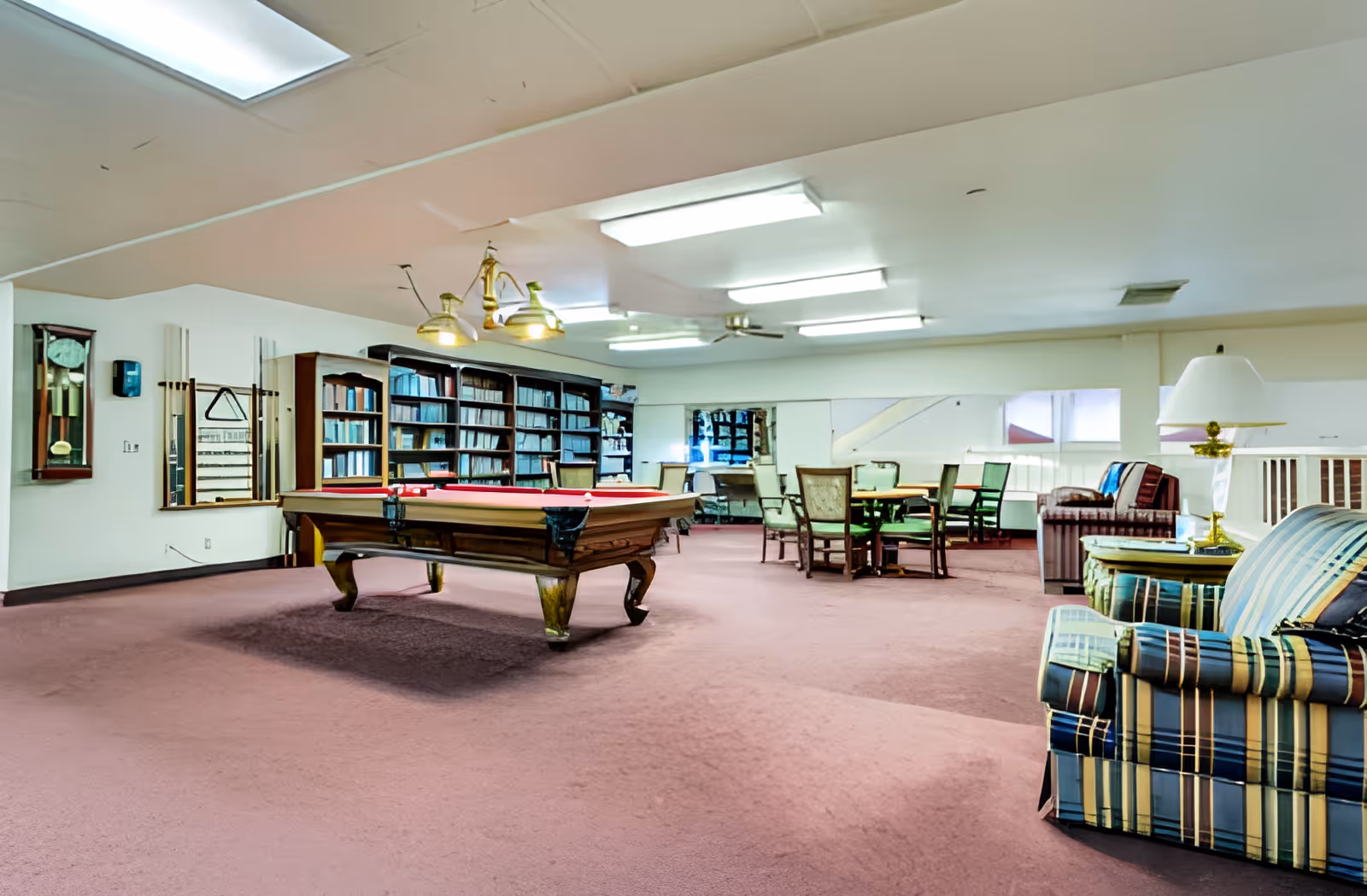 A spacious recreational room with a pool table in the center, surrounded by bookshelves filled with books. There are several chairs and tables for seating, along with a striped armchair and a lamp on a side table. The room has a carpeted floor and fluorescent ceiling lights.