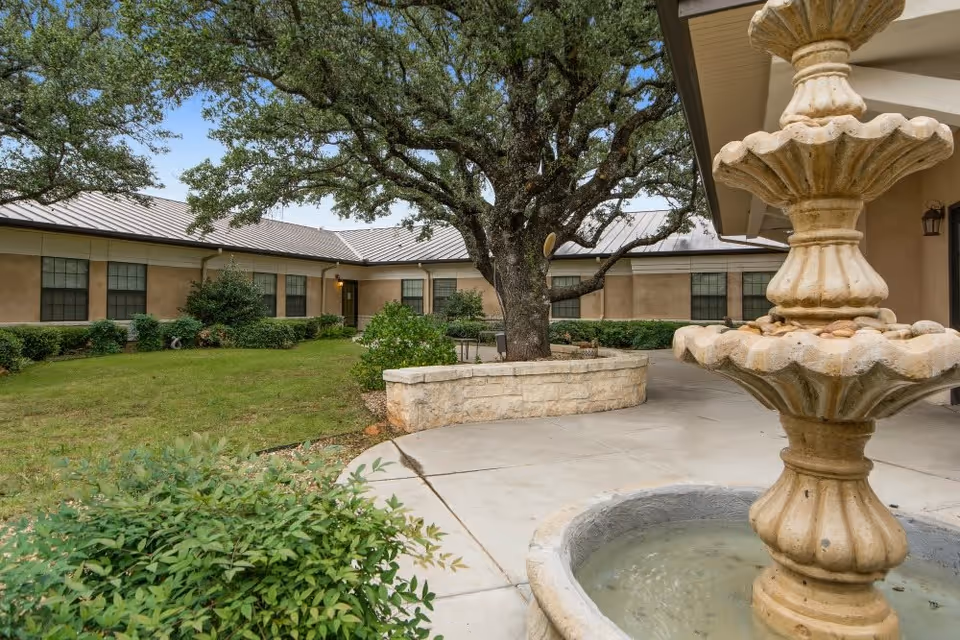 Outdoor courtyard area of Arbor House Assisted Living and Memory Care - Llano featuring a large tree surrounded by a stone planter, a multi-tiered stone water fountain in the foreground, green bushes, and a beige building with multiple windows in the background under a clear sky.