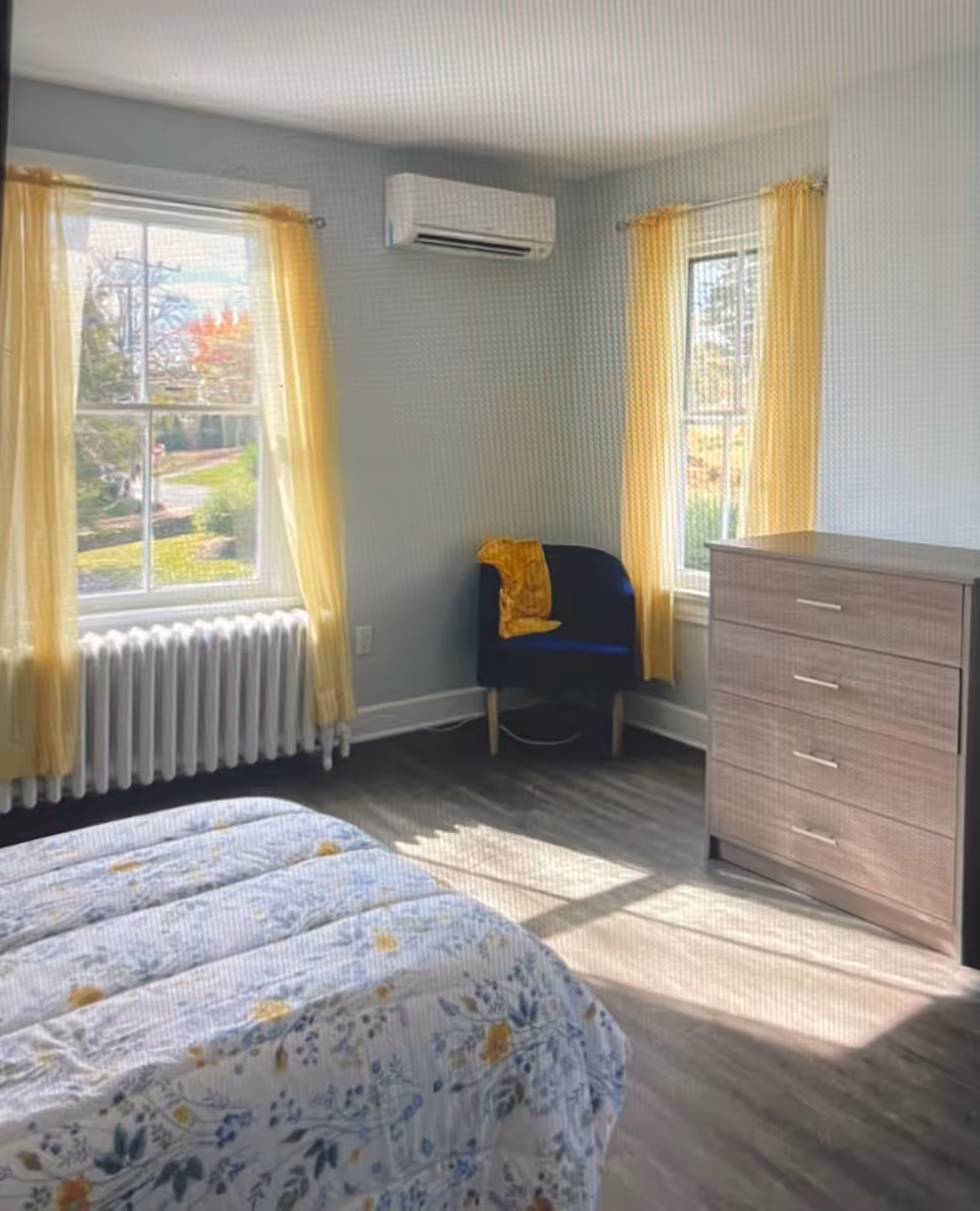 A bright bedroom with two windows covered by yellow curtains, a bed with floral bedding, a blue chair with a yellow blanket draped over it, a wooden dresser with four drawers, and a wall-mounted air conditioning unit.