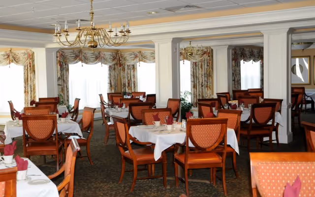 A formal dining room with multiple round and rectangular tables covered with white tablecloths. Each table is set with cups, plates, and folded red napkins. The chairs have wooden frames with orange patterned upholstery. The room features large windows with floral curtains, white columns, a patterned carpet, and a brass chandelier hanging from the ceiling.