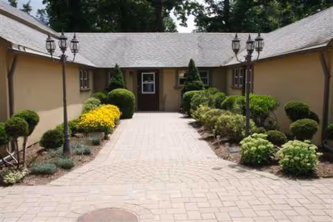 Pathway lined with trimmed shrubs leads to a central door between beige buildings under lamp posts