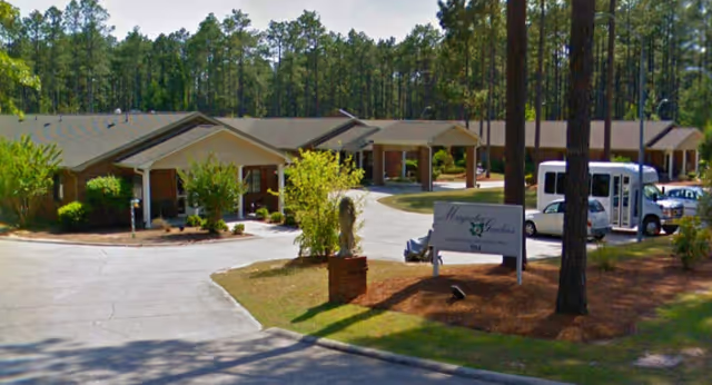 Exterior view of Magnolia Gardens senior living facility showing single-story buildings surrounded by tall pine trees, a driveway, parked vehicles including a white shuttle bus, and a sign with the facility's name at the entrance.