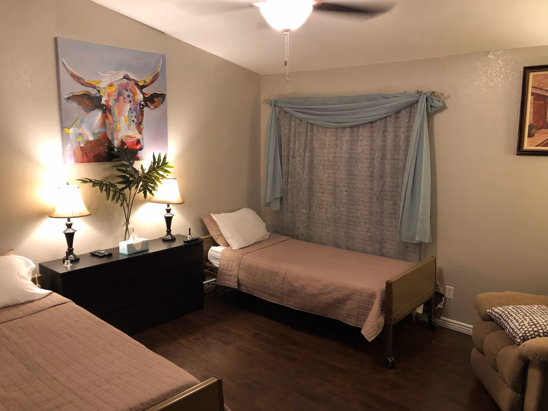 Cozy bedroom with two single beds, a black dresser topped with lamps and a vase, and a colorful cow painting on the wall.