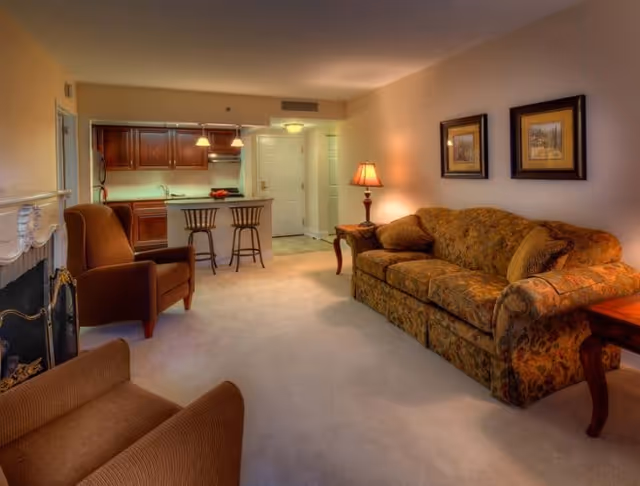 A cozy living room with a patterned sofa, two armchairs, a side table with a lamp, and two framed pictures on the wall. The room opens into a kitchen area with wooden cabinets, a countertop with two bar stools, and a white door in the background.