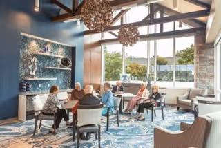 A group of elderly people sitting around a table in a bright, spacious common area with large windows, modern decor, and blue patterned carpet. The room features wooden beams on the ceiling, decorative hanging light fixtures, and shelves with artistic objects.
