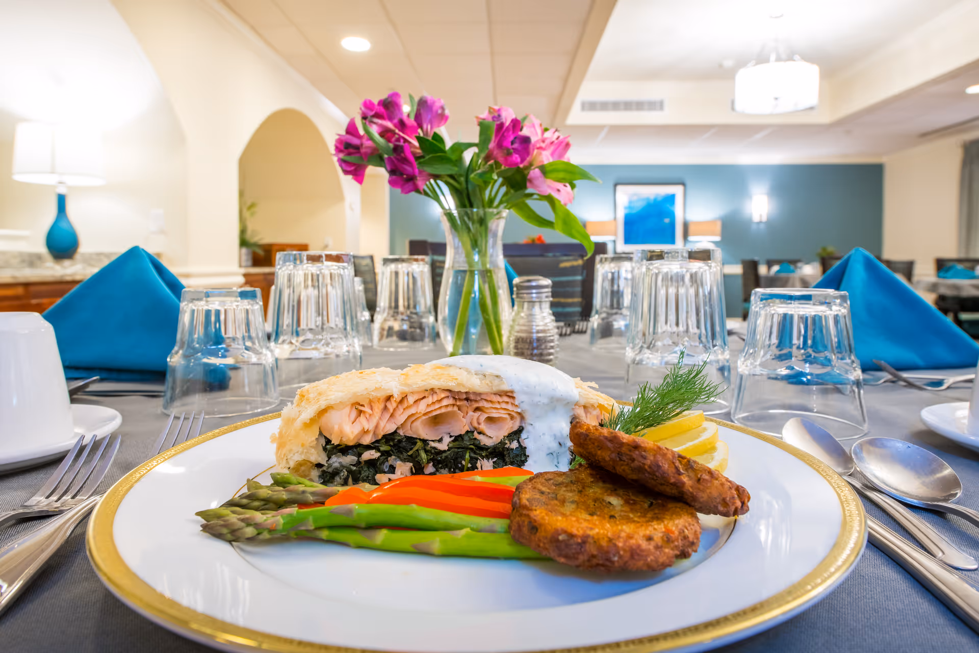 A plated meal featuring a salmon fillet wrapped in pastry with a creamy sauce, served with asparagus, red bell pepper strips, two potato cakes, and lemon slices garnished with dill. The plate is set on a table in a dining room with blue napkins, upside-down glasses, silverware, and a vase of pink flowers in the background.