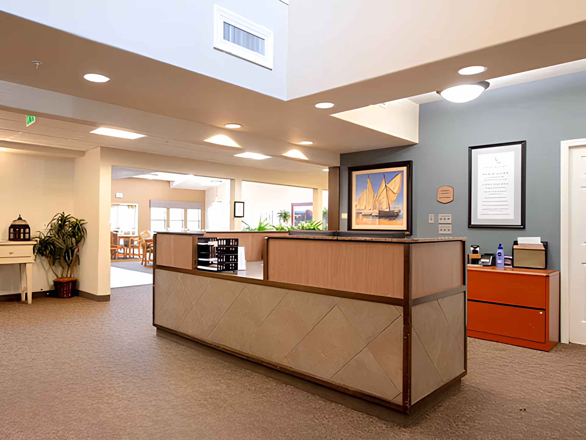Reception area inside a senior living facility with a wooden front desk, beige carpet, and a blue accent wall. The desk has office supplies on it, and there is a framed picture of sailboats and a framed document on the wall behind the desk. In the background, there is a dining area with tables and chairs, and some plants are placed around the space.