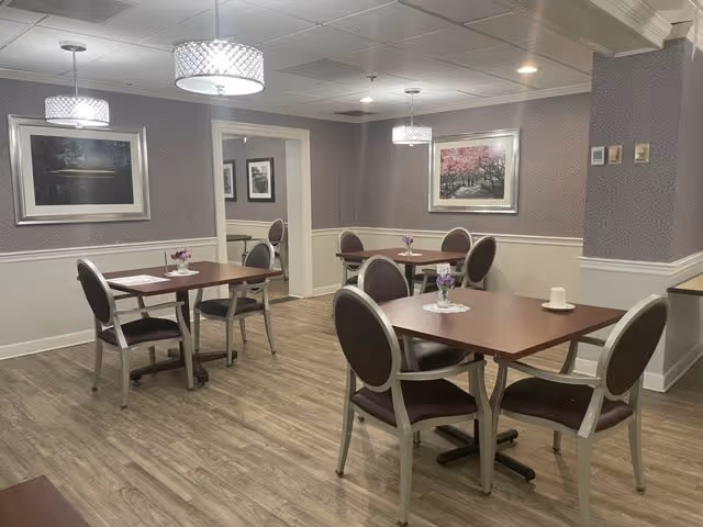 A dining room in a senior living community with four wooden tables, each surrounded by four chairs with dark cushioned seats and backs. The room has wood flooring, patterned wallpaper, and framed artwork on the walls. Modern ceiling lights hang above the tables, and small flower arrangements are placed on each table.