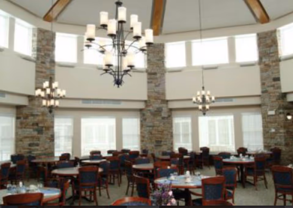 Spacious dining room with multiple round tables and wooden chairs with blue upholstery, large windows letting in natural light, stone pillars, and elegant chandeliers hanging from a high ceiling with wooden beams.