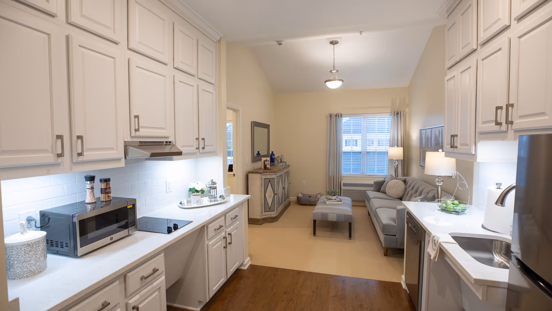 A bright and modern kitchen area with white cabinets, a microwave, stovetop, and sink on either side. The kitchen opens into a cozy living room with a gray sofa, ottoman, side table with a lamp, and a window with curtains letting in natural light.
