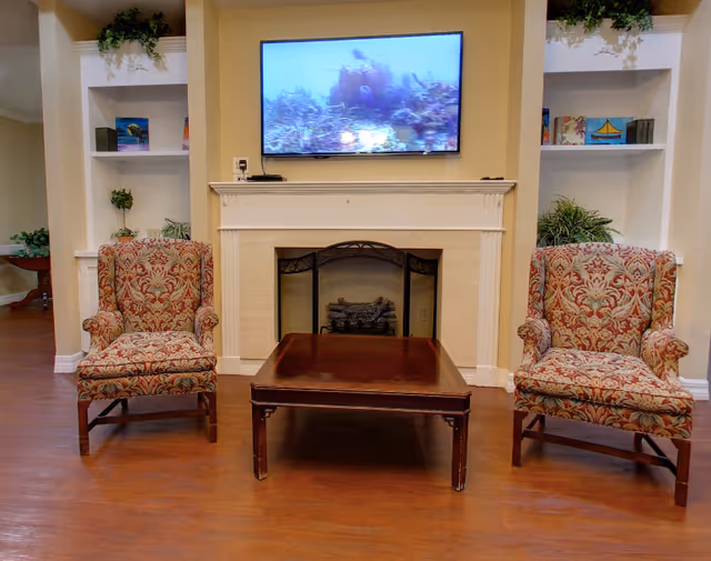 Two patterned armchairs flank a wooden coffee table in front of a fireplace with a TV mounted above in a communal living room.