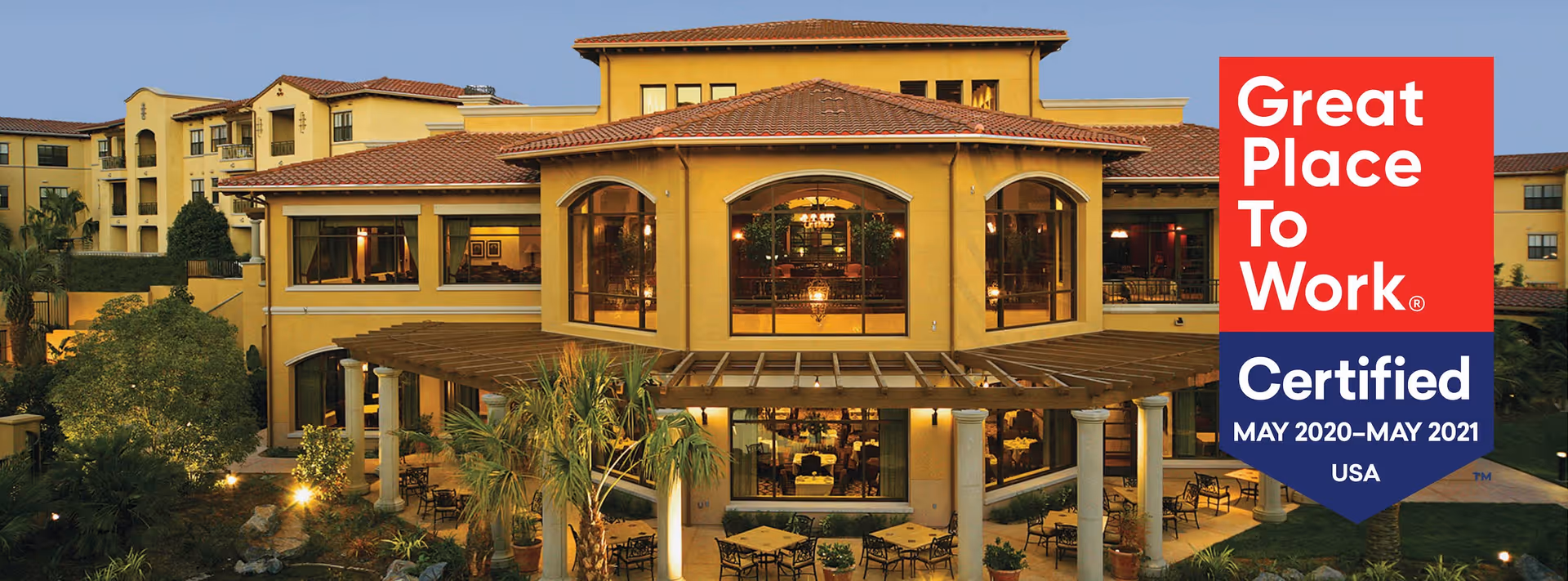 Front exterior of a Mediterranean-style senior living building with a covered patio and outdoor seating, shown with a certification badge.