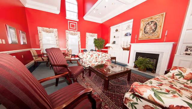 A bright living room with red walls and white trim featuring floral patterned sofas, striped armchairs, a glass coffee table on a patterned rug, a fireplace with a plant inside, built-in white shelves, and large windows with floral curtains.
