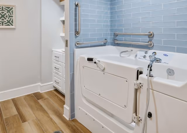 A bathroom featuring a walk-in bathtub with safety grab bars on the walls. The bathtub has a door for easy access and a handheld showerhead. The walls are tiled with light blue subway tiles, and the floor has wood-look tiles. A white vanity with drawers is partially visible in an adjacent area.