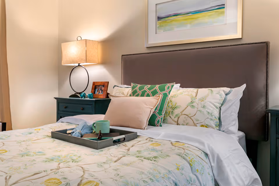 A well-appointed bedroom with a made bed featuring floral bedding and decorative pillows, a tray on the bed, and a nightstand with a lamp and framed photo.