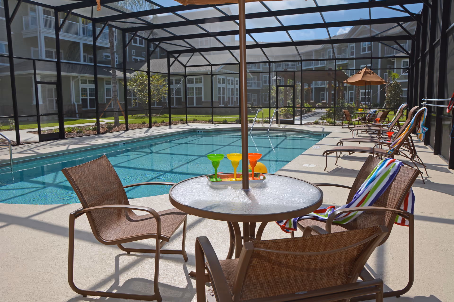 A screened-in outdoor pool area with lounge chairs and a round glass table with an umbrella. Colorful towels are draped over some chairs, and three colorful cocktail glasses are on the table. The pool is surrounded by a beige deck, and residential buildings are visible in the background.