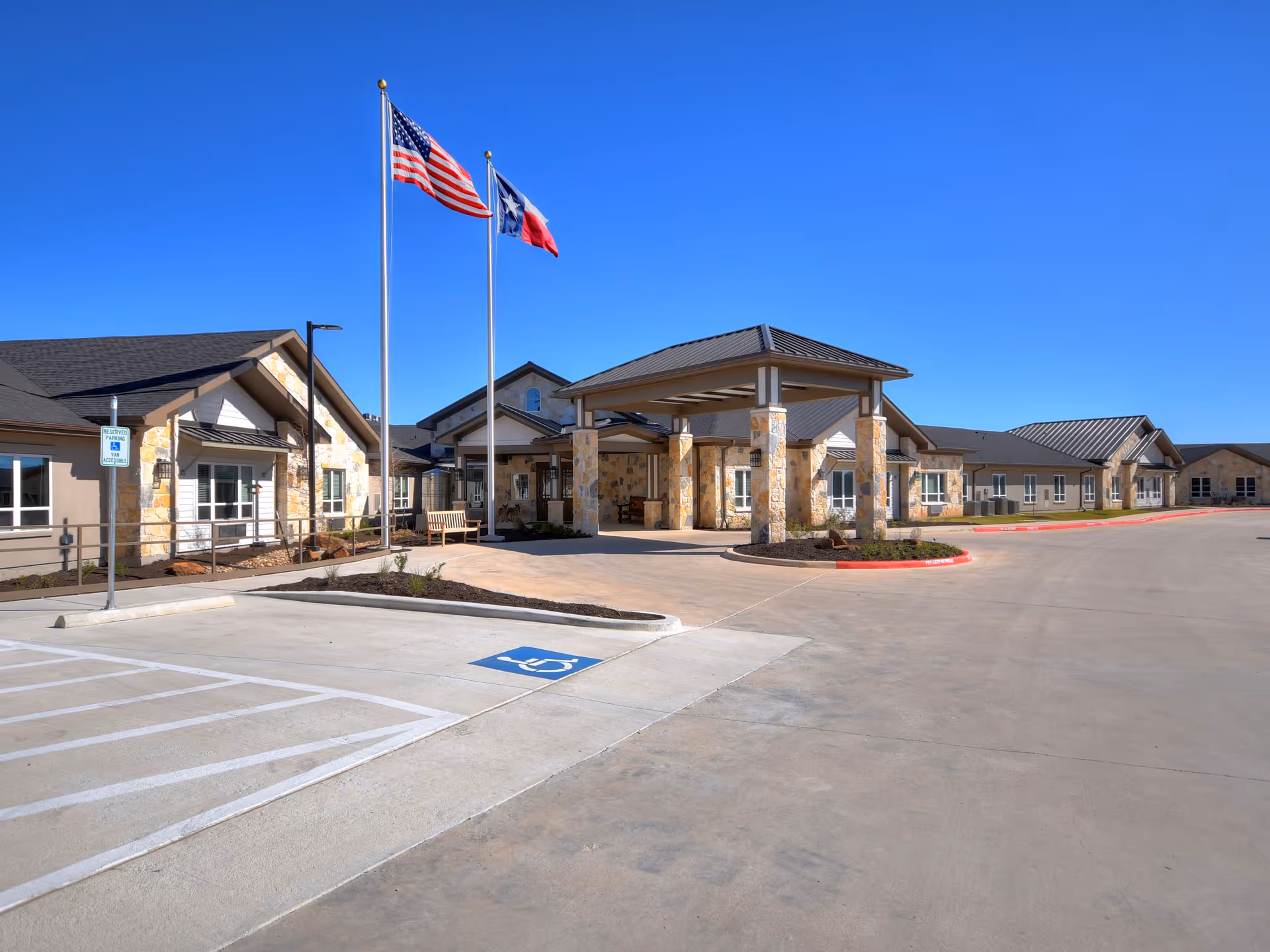 Exterior view of Alexis Pointe of Wimberley Senior Living facility showing a single-story building with stone and beige walls, a covered entrance supported by stone pillars, two flagpoles with the American and Texas flags, and a parking area with a handicapped parking space in front.