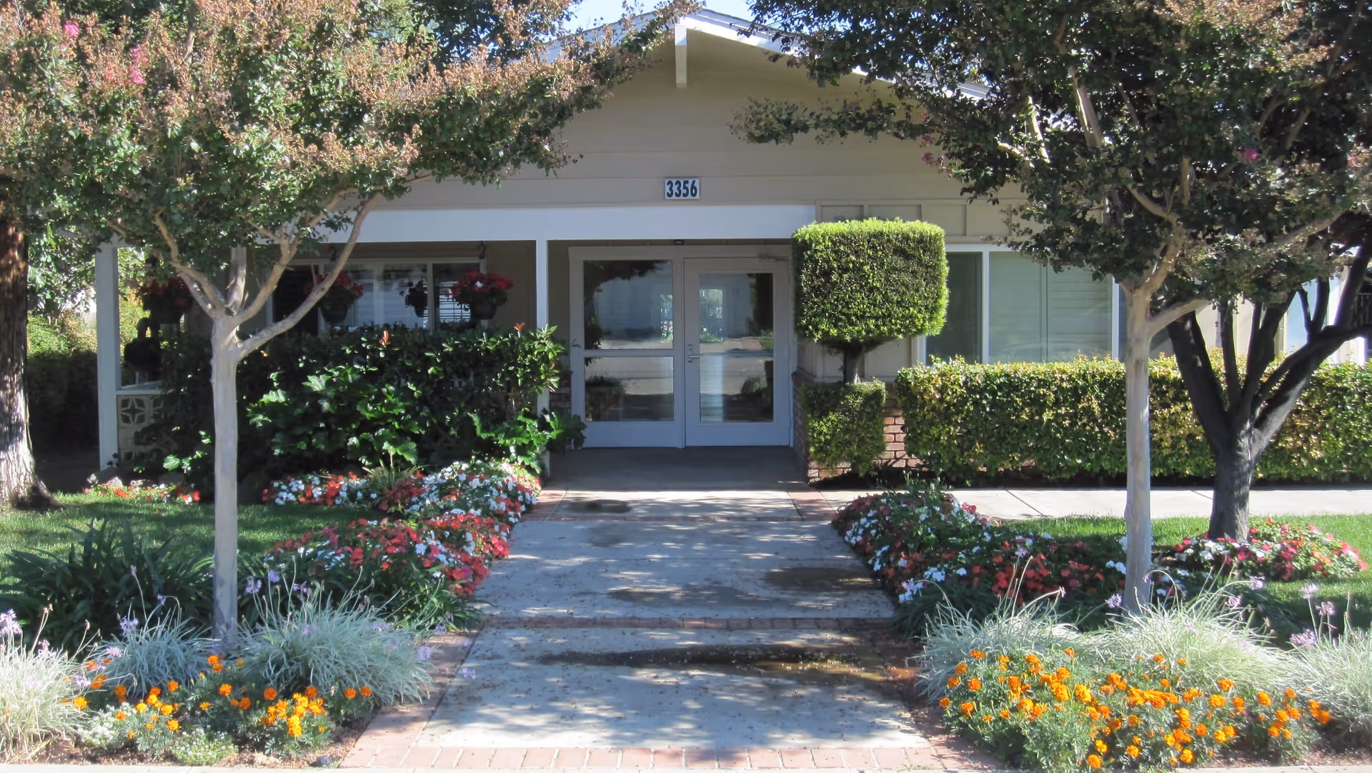 Front entrance of a single-story senior living building with a walkway, flowering landscaping, and double glass doors.