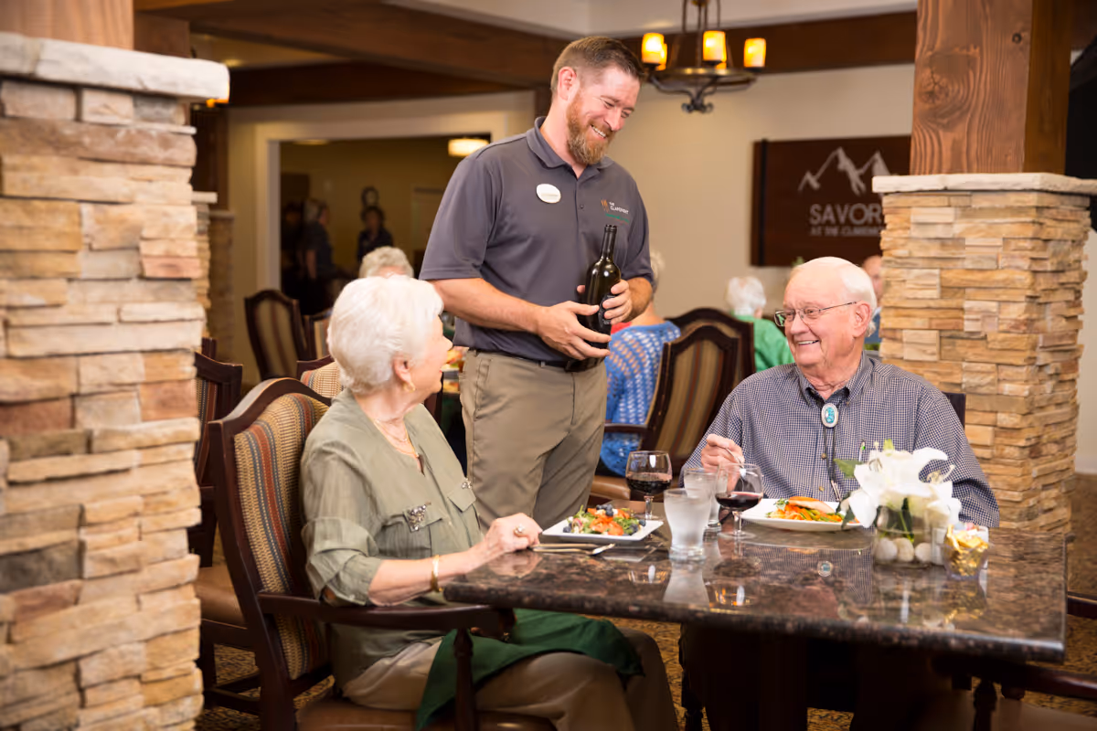 A server pours wine for two older adults seated at a table in a dining room with plates of food.