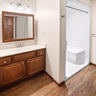 Bathroom with wooden floor, wooden vanity with white countertop and sink, a large mirror with wooden frame above the sink, three-light fixture above the mirror, and a white shower area with built-in seat and a shower curtain rod.